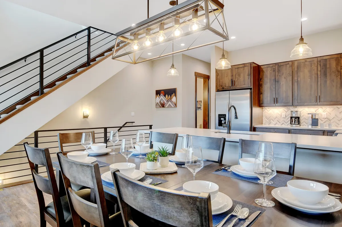 A dining table set for a meal sits beneath an industrial-style light fixture, with the adjoining modern kitchen cabinetry in view. The scene is warm and inviting, clearly designed for entertaining and group meals.