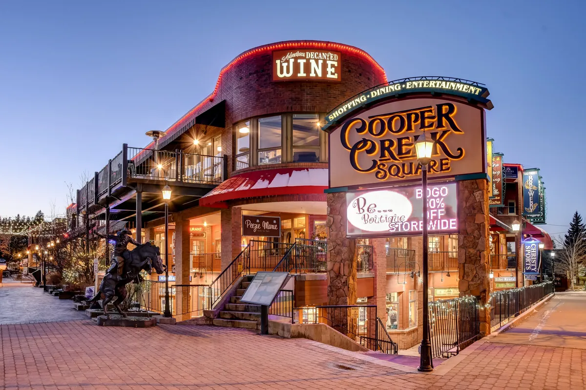 A vibrant exterior shot of Cooper Creek Square, featuring the recognizable square sign, a wine shop entrance, festive decorative lighting, and a modern sculpture in the foreground. People are seen strolling, hinting at a lively, walkable nightlife and dining scene.