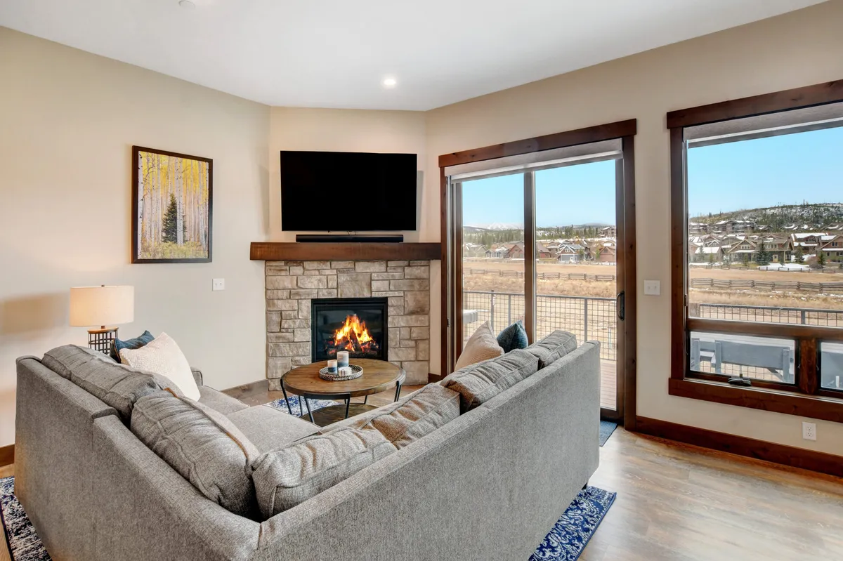 This image features a welcoming living room anchored by a modern fireplace, a plush sofa, a large coffee table, and expansive windows that look out onto mountain views. The space is bathed in natural light and has a cozy atmosphere ideal for gathering and relaxing.